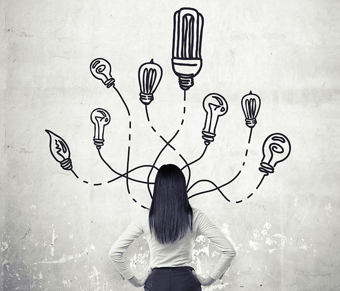 A woman with hands on hips standing in front of a concrete wall with many different bulbs drawn around her head. Back view. Concrete background. Concept of having an idea.